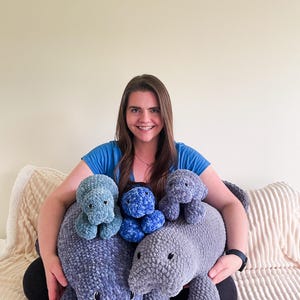 Puede incluir: Una mujer est&aacute; sentada en un sof&aacute; y sostiene cuatro peluches de manat&iacute; tejidos a crochet. Los peluches son de varios tonos de azul y gris.