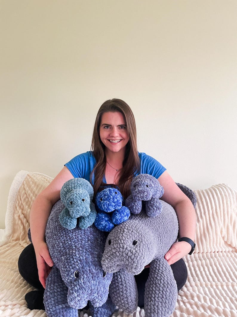 Puede incluir: Una mujer est&aacute; sentada en un sof&aacute; y sostiene cuatro peluches de manat&iacute; tejidos a crochet. Los peluches son de varios tonos de azul y gris.