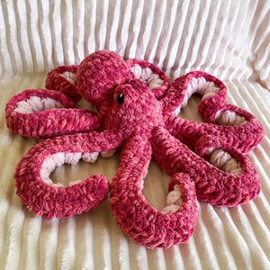 Può includere: Un polpo di peluche fatto a mano, in tonalità di rosa. Il polpo ha una testa rotonda, otto tentacoli arricciati e piccoli occhi neri. Il lato inferiore dei tentacoli è di un rosa più chiaro. Il giocattolo è realizzato in un materiale morbido e strutturato.