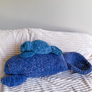 Puede incluir: Dos peluches de manat&iacute; azul hechos a crochet. El manat&iacute; m&aacute;s grande est&aacute; acostado boca abajo con las aletas extendidas. El manat&iacute; m&aacute;s peque&ntilde;o est&aacute; sentado sobre la espalda del m&aacute;s grande.