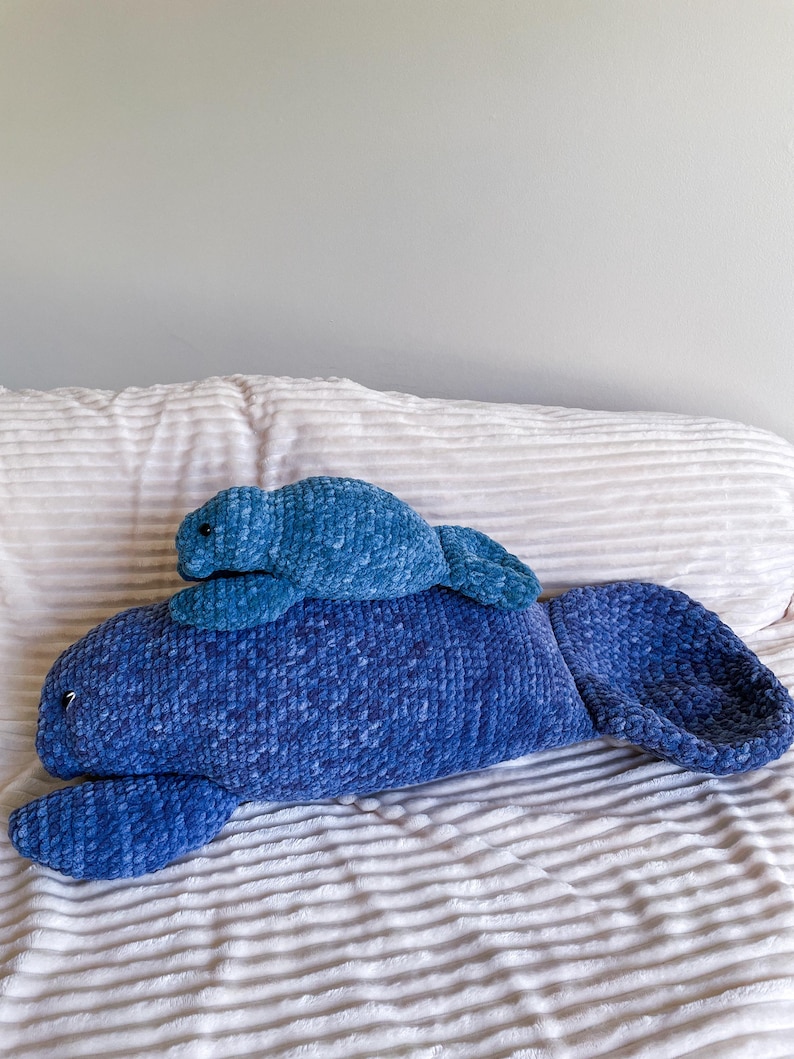 Puede incluir: Dos peluches de manat&iacute; azul hechos a crochet. El manat&iacute; m&aacute;s grande est&aacute; acostado boca abajo con las aletas extendidas. El manat&iacute; m&aacute;s peque&ntilde;o est&aacute; sentado sobre la espalda del m&aacute;s grande.