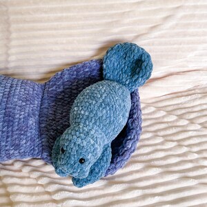 Puede incluir: Un juguete de peluche azul de crochet con forma de manat&iacute; est&aacute; acostado sobre una manta blanca y esponjosa. El juguete tiene una cola peque&ntilde;a y aletas grandes.