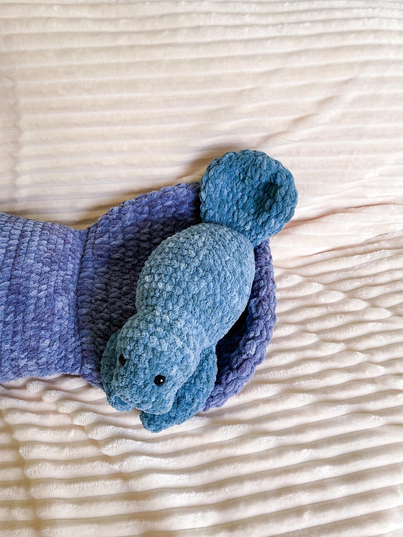 Puede incluir: Un juguete de peluche azul de crochet con forma de manat&iacute; est&aacute; acostado sobre una manta blanca y esponjosa. El juguete tiene una cola peque&ntilde;a y aletas grandes.