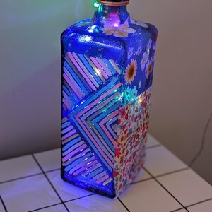 Könnte beinhalten: Eine blaue Glasflasche mit einem Korkverschluss und einem floralen und geometrischen Design. Die Flasche wird von innen mit bunten Lichterketten beleuchtet.