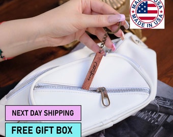 May include: White faux leather fanny pack with a zipper closure and a silver zipper pull. A wooden keychain with the text "Christmas" is attached to the zipper pull. The keychain is hanging out of the fanny pack. The fanny pack is on a wooden surface. The text "NEXT DAY SHIPPING" and "FREE GIFT BOX" are on pink and blue rectangles, respectively, in the lower left corner of the image.