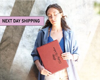 May include: A red leather-bound journal with the name "Ashley Watson" engraved in gold lettering. The journal has a floral design and is held by a person wearing a blue shirt.