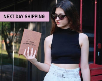 May include: A brown leather journal with the name "Lauren" engraved on the front. The journal is held by a person wearing a black sleeveless top and white pants.