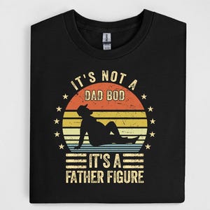 May include: Black t-shirt with a graphic design. The design features a silhouette of a person reclining, set against a sunset-style background with the text "IT'S NOT A DAD BOD IT'S A FATHER FIGURE".