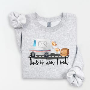 May include: A light gray sweatshirt featuring a watercolor illustration of a mail truck with a package and the phrase "this is how I roll" in black script. The mail truck is white with blue and red accents.