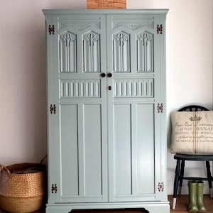 May include: A tall, light blue painted wooden wardrobe featuring two doors with intricate carved panels, vertical moulding, and dark metal hinges. The cabinet has two small round knobs and a central keyhole. A wooden box labelled BEUREL sits on top.