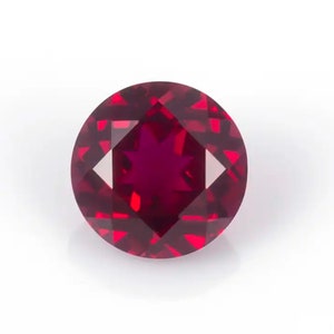 May include: A round, faceted ruby gemstone. The gem is a deep red color with a star-like pattern in the center. The facets create a brilliant sparkle, and the stone is set against a white background.