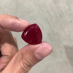 Könnte beinhalten: Ein tief roter Edelstein mit dreieckiger Form, in Facetten geschliffen. Der Edelstein wird zwischen zwei Fingern gehalten.