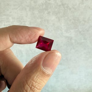 Puede incluir: Una gema de color rojo intenso y vibrante tallada en forma rectangular con superficies facetadas que reflejan la luz. La piedra muestra un tono carmesí rico y saturado, con una calidad transparente y clara y bordes geométricos precisos.