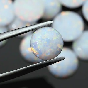 May include: A close-up of a white opal cabochon, held by tweezers. The opal has a milky white base with iridescent flashes of color.