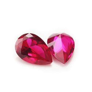 Może przedstawiać: Dwa fasetowane, gruszkowate kamienie szlachetne w żywym odcieniu magenty. Kamienie są prezentowane na białym tle, ukazując ich brylantowy szlif i odblaskowe powierzchnie. Te kamienie szlachetne są prawdopodobnie przeznaczone do tworzenia biżuterii lub kolekcjonowania.