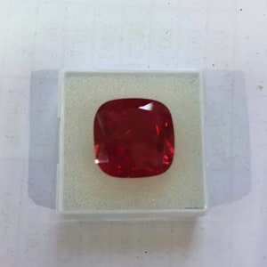 May include: A deep red gemstone with a cushion cut, displayed in a white box.