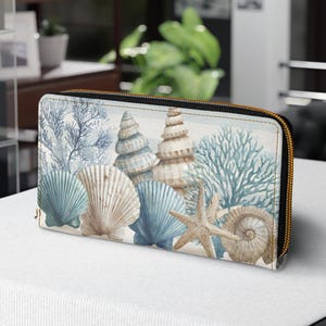 May include: A zippered wallet featuring a coastal design with seashells, starfish, and coral in shades of blue, beige, and white. The wallet has a gold-colored zipper and a rectangular shape.