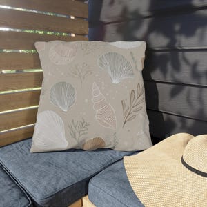 Pode incluir: Uma almofada quadrada bege com um padrão de conchas em branco, castanho e cinza. A almofada está sobre uma almofada azul, com um chapéu de palha visível. O design apresenta várias ilustrações de conchas.