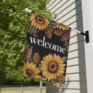 Puede incluir: Una bandera decorativa para exteriores con un fondo negro y la palabra "welcome" en blanco. La bandera está adornada con girasoles amarillos y piñas marrones. La bandera está montada en un poste blanco y negro.