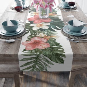 May include: A white table runner with a watercolor floral design featuring pink hibiscus, white plumeria, and green tropical leaves. The runner is set on a wooden table with place settings and a vase of pink lilies.