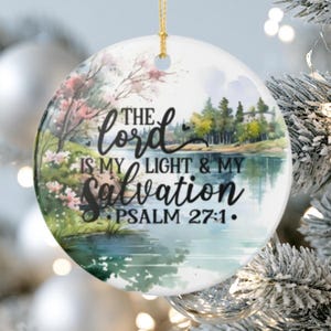 Peut inclure: Une décoration ronde en céramique blanche ornée d'un paysage à l'aquarelle représentant un lac, des arbres et des fleurs roses. Le texte noir indique : The Lord is my light & my salvation Psalm 27:1. Un cordon doré est fixé au sommet.