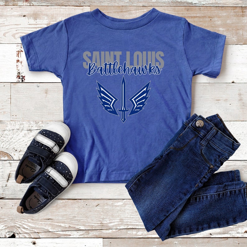 Puede incluir: Una camiseta azul con el texto "Saint Louis Battlehawks" en gris y un gr&aacute;fico de espada azul con alas.
