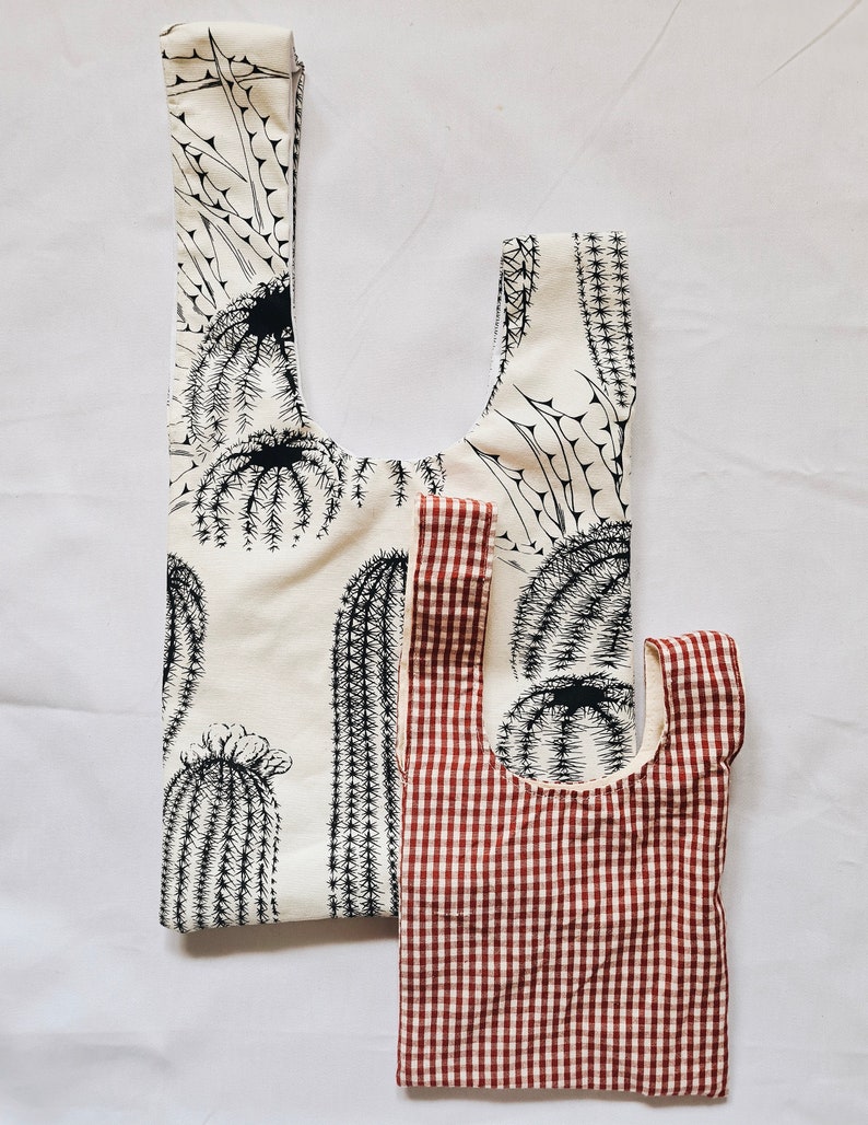 May include: Two reusable shopping bags. One is white with a black cactus print. The other is red and white gingham.