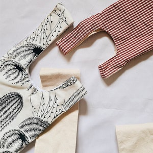 May include: Three reusable shopping bags. One bag is white with a black cactus print. Another bag is white with a red gingham pattern. The third bag is plain white.