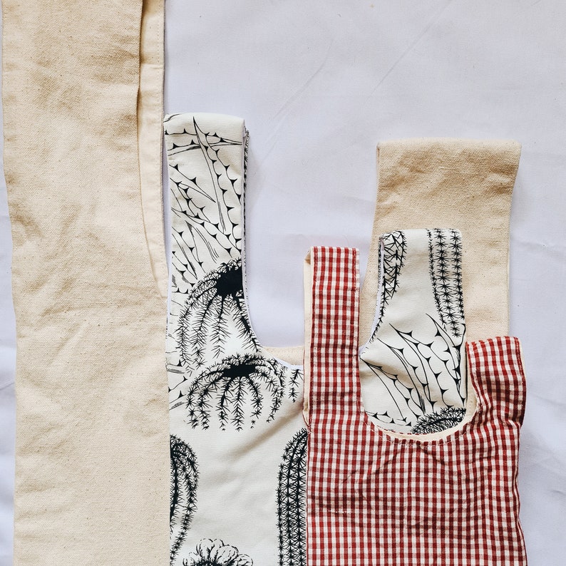 May include: Three reusable shopping bags made of natural fabric. One bag is white with a black cactus print, another is white with a red gingham pattern, and the third is a plain white bag.