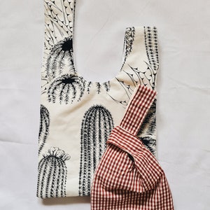 May include: Two reusable shopping bags. One is white with a black cactus print. The other is red and white gingham.