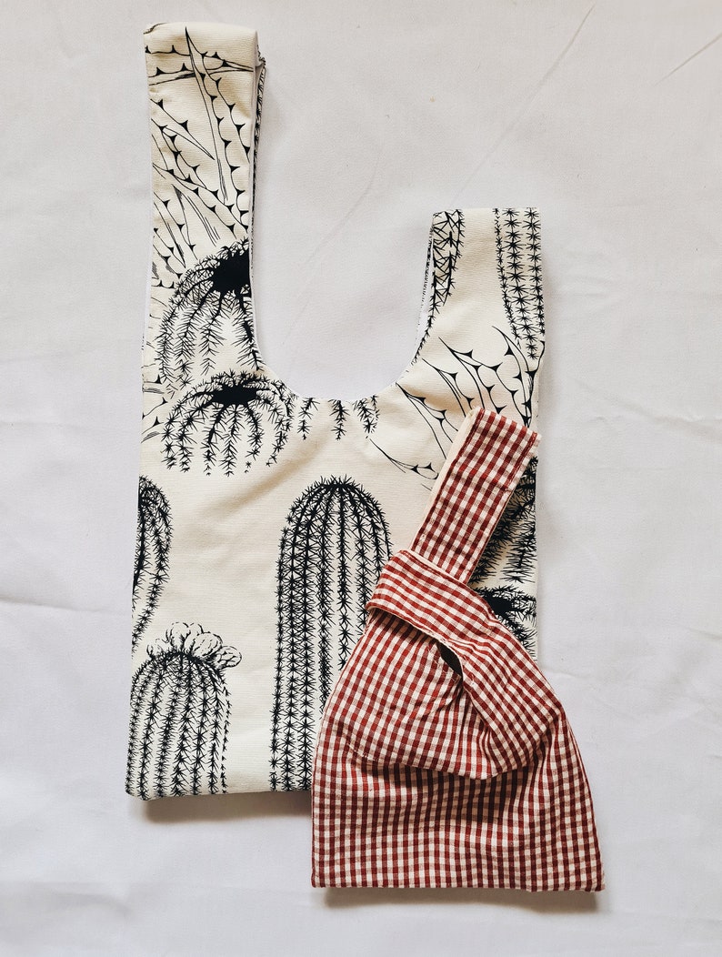 May include: Two reusable shopping bags. One is white with a black cactus print. The other is red and white gingham.