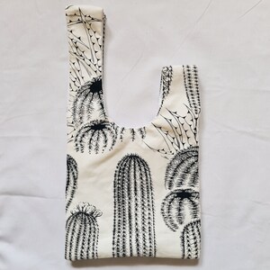 May include: A white tote bag with a black cactus print. The bag has a wide, flat base and a single handle.