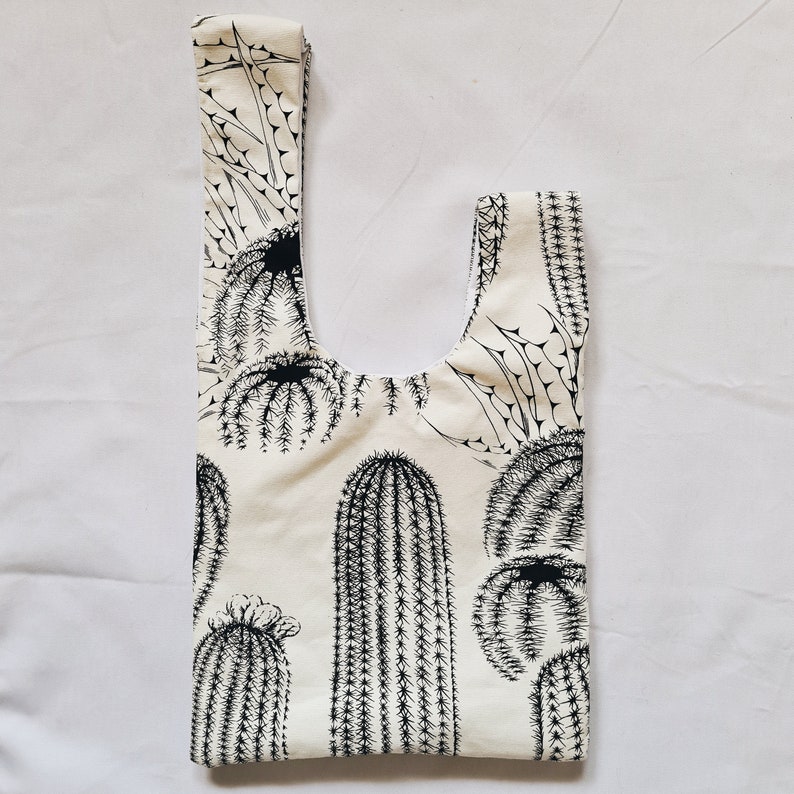 May include: A white tote bag with a black cactus print. The bag has a wide, flat base and a single handle.