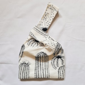 May include: A white fabric tote bag with a black cactus print. The bag has a knotted handle and is made of a soft, lightweight material.