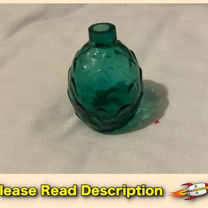 May include: A small, translucent emerald green glass bottle with a rounded, bulbous body and a textured, repeating scale-like pattern across its surface. The bottle features a short, narrow neck with a circular opening at the top.