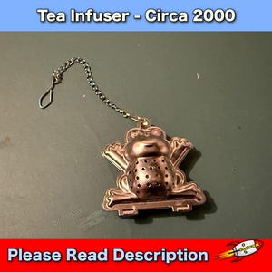 May include: A metal tea infuser shaped like a frog, with a chain and a hook. The infuser has a perforated body for steeping tea leaves. The text "Tea Infuser - Circa 2000" is at the top. The text "Please Read Description" is at the bottom.