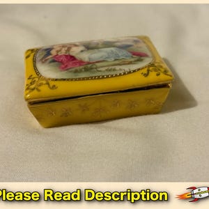 May include: A rectangular yellow porcelain trinket box featuring a central painted scene of figures in colorful clothing. The lid is bordered by gold filigree accents, and the sides of the box are decorated with a delicate gold floral pattern.