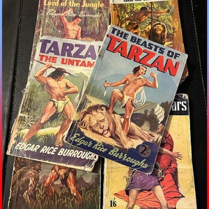 May include: A stack of vintage paperback books featuring Tarzan, the jungle hero, by Edgar Rice Burroughs. The covers feature illustrations of Tarzan, a lion, and other jungle scenes. The books are priced at 2/6, 2/-, and 1/6.