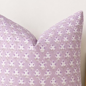 May include: A square throw pillow featuring a light lavender fabric base with a repeating pattern of small, stylized white and purple floral motifs arranged in a grid. The pillow has a soft, matte texture and a plump, filled appearance.