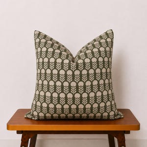 May include: A decorative throw pillow with a green background and a repeating pattern of white floral shapes. The pillow is placed on a small wooden bench with a warm, brown finish. The pillow's design adds a touch of nature-inspired style.