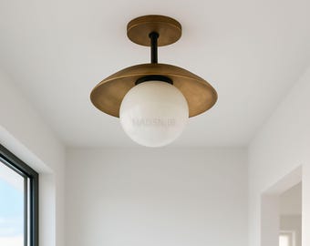 May include: A brass-toned ceiling light fixture with a white globe-shaped shade. The fixture has a circular base and a wide, disc-shaped shade. The text "MADSNIJB" is printed on the globe. The light is mounted on a white ceiling.
