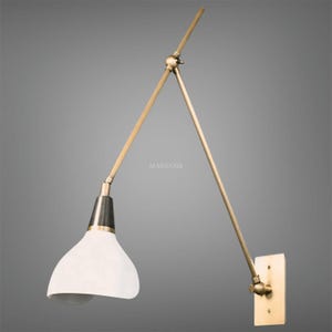 May include: A wall-mounted brass lamp with a white shade. The lamp has an adjustable arm and a rectangular base. The design is modern and minimalist, suitable for various interior styles. The lamp's arm is adjustable for directional lighting.