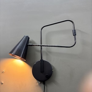 May include: A black wall-mounted lamp with a cone-shaped shade and a rectangular arm. The lamp is mounted on a circular base with a power cord. The light is on, illuminating the interior of the shade.