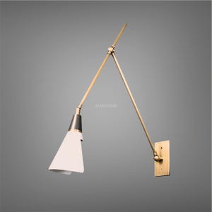 May include: A contemporary wall lamp with a brass finish. It has a white, cone-shaped shade and an adjustable arm. The lamp is mounted on a rectangular brass plate. The text "MADSNIB" is visible.