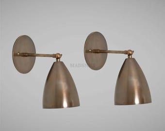 May include: Two bronze-colored wall sconces. Each sconce features a circular backplate, a straight arm, and a conical shade. The fixtures are made of metal and have a vintage or industrial aesthetic. The sconces are mounted on a light gray wall.