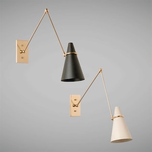 Op de afbeelding: Twee wandlampen in een mid-century modern design. Eén lamp heeft een zwarte conische kap, de andere een gebroken witte kap. Beide hebben een goudkleurige verstelbare arm en een rechthoekige goudkleurige wandbevestiging.