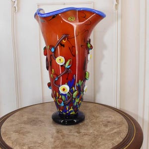 Może przedstawiać: Wysoki, pomarańczowy wazon ze szkła artystycznego z rozszerzaną krawędzią i czarną podstawą. Wazon jest ozdobiony kolorowymi kwiatowymi i liściastymi akcentami. Obrzeże jest wykończone na niebiesko. Wazon jest wystawiony na okrągłym stole z marmurowym blatem.