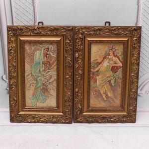 May include: A pair of framed art prints with Art Nouveau style illustrations of women in flowing gowns. The gold-coloured frames are ornate and rectangular. The artwork depicts figures in shades of green, red, and gold.