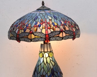 Puede incluir: Una lámpara de mesa de vidrio Tiffany con un diseño de libélula. La lámpara tiene una pantalla de vidrio Tiffany azul, verde y roja y una base a juego. La lámpara está encendida y la luz brilla a través del vidrio Tiffany.