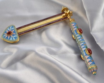 May include: An ornate, gold-toned decorative object with a blue, red, and gold design. The object has a triangular end with a red stone and a cylindrical handle with red stones. The background is a silver satin fabric.